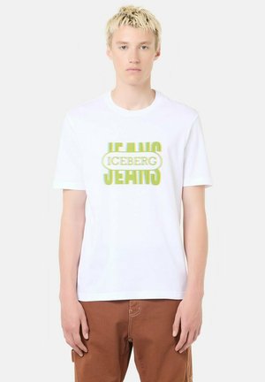 Giovane con capelli biondi corti che indossa una maglietta bianca con scritta giallo-verde "ICEBERG JEANS" e pantaloni marroni, in piedi davanti a uno sfondo semplice.
