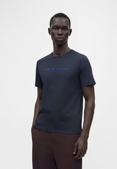 Άνδρας μοντέλο που φοράει μπλε σκούρο t-shirt Tiger of Sweden και καφέ παντελόνια, στέκεται μπροστά σε απλό ανοιχτό γκρι φόντο.