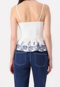 Haut blanc smocké avec fines bretelles, présentant un motif floral bleu à l'ourlet. Associé à un jean bleu foncé, offrant une coupe décontractée.