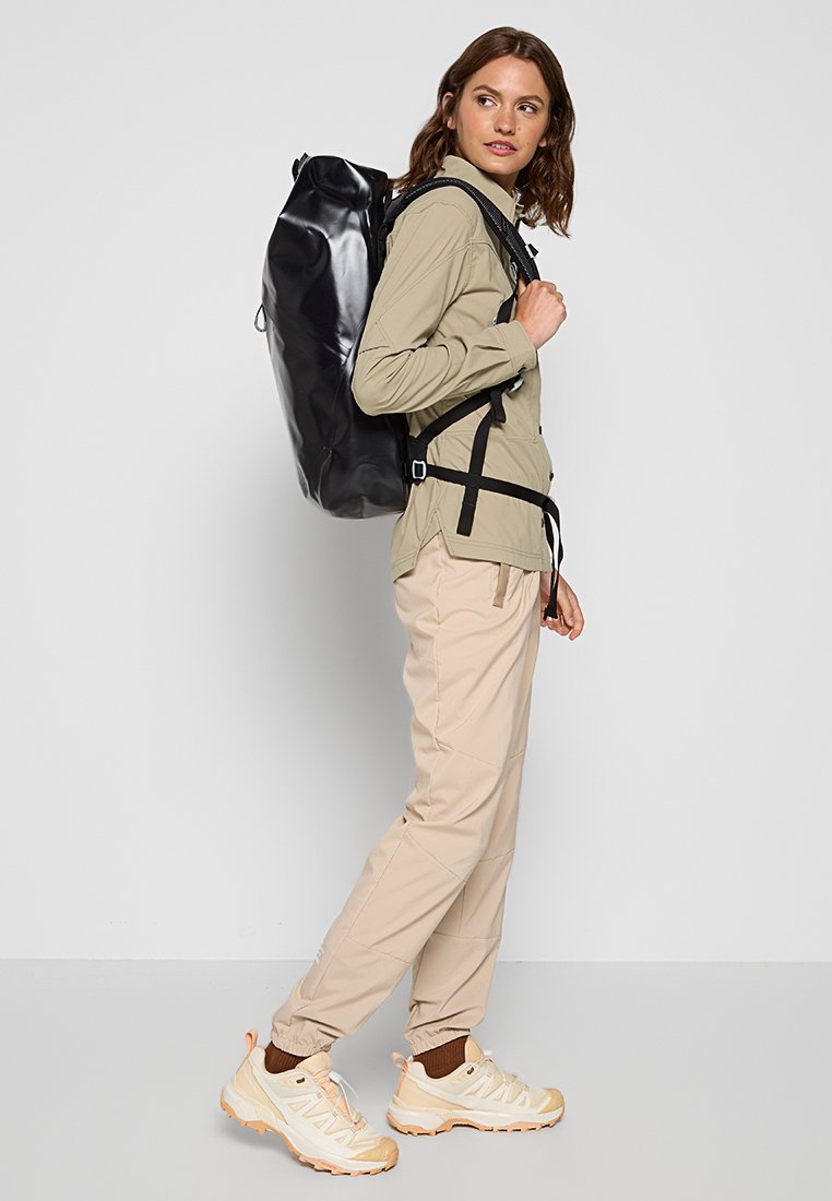 Sac à dos noir imperméable avec un dessus roulé, des bretelles réglables et un design épuré. Porté avec une chemise à manches longues beige clair et un pantalon.