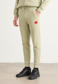 Light green joggers with an elastic waistband, drawstring, side pockets, and a red rectangular logo patch on the thigh. Black sneakers included.