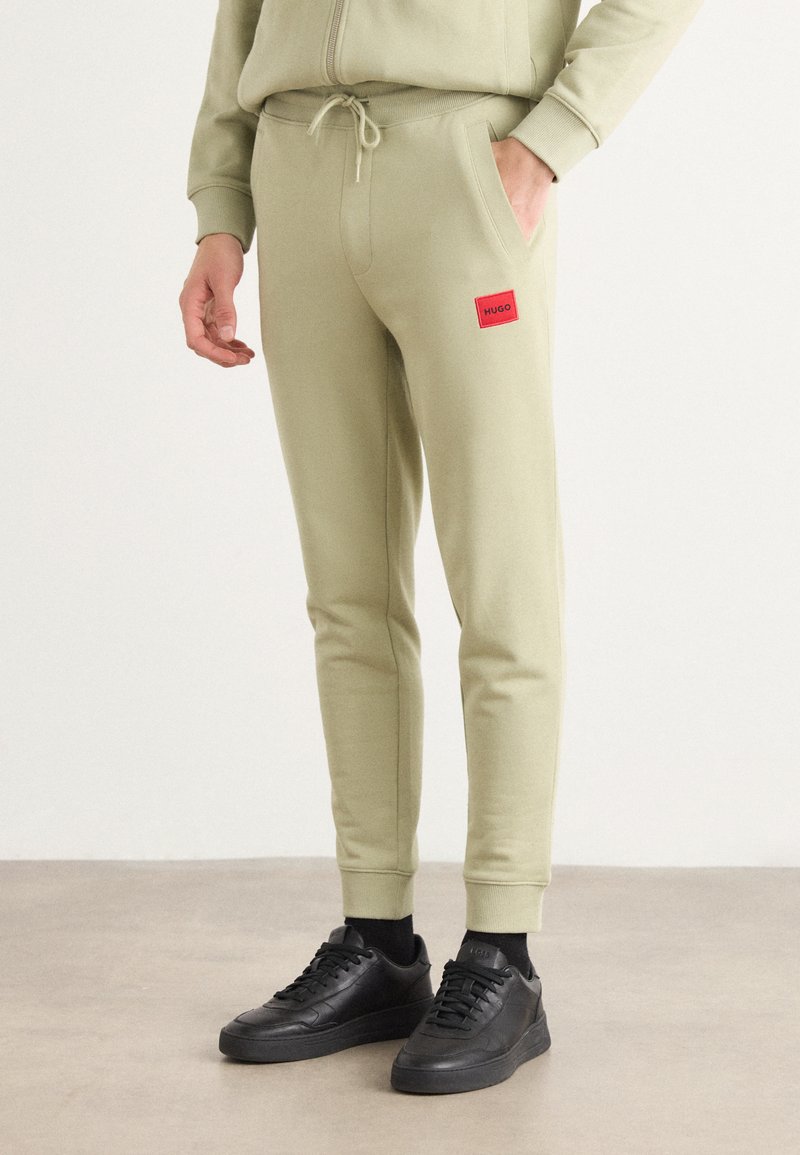 Light green joggers with an elastic waistband, drawstring, side pockets, and a red rectangular logo patch on the thigh. Black sneakers included.
