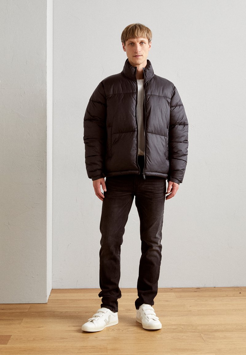 Doudoune noire avec un col montant, design matelassé et fermeture éclair. Portée par-dessus une chemise grise avec un jean foncé et des baskets blanches.