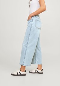 Kvinna som bär ljusblå vida jeans, vita sneakers med svarta detaljer, och en vit kortärmad topp, stående mot en enfärgad bakgrund.