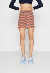 Pantaloni corti con motivo zigzag multicolore in rosso, rosa e marrone, con orlo svasato. Indossati con scarpe con tacco a piattaforma a strappi di colore blu chiaro.