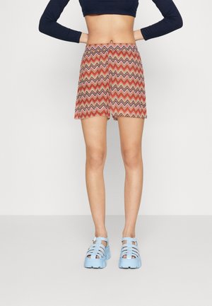 Shorts à motif zigzag multicolore en rouge, rose et marron avec un ourlet évasé. Portés avec des chaussures à plateforme à lanières bleu clair.