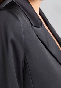 Schwarzer Satin-Blazer mit Reverskragen, der eine glatte Textur und eine leicht strukturierte Passform bietet. Die Seitenansicht hebt die Reversdetails und den Glanz des Stoffes hervor.