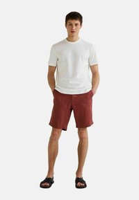 T-shirt en coton blanc, shorts en lin rouges et sandales noires ; une tenue décontractée mettant en avant un design simple et une coupe relaxe.