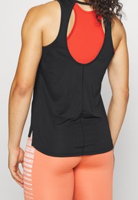 Zwarte mouwloze tanktop met een open rug, gelaagd over een rode sportbeha. De stof lijkt glad en heeft subtiele stiksels.