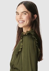 Oliwkowa sukienka z falbankami wzdłuż dekoltu i rękawów, o miękkiej, swobodnej teksturze. Modelka spogląda lekko do przodu.