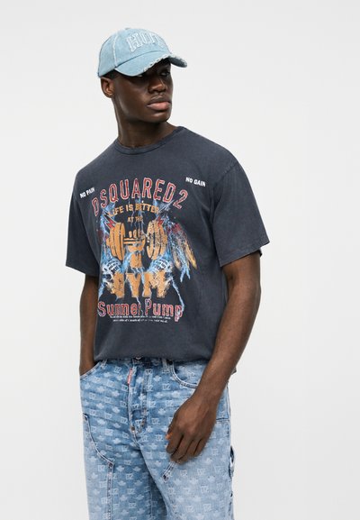 Άνδρας που φοράει μπλε σκουριασμένο καπέλο, μαύρο γραφικό T-shirt με την επιγραφή "DSQUARED2" και τζιν με λευκό μοτίβο λογοτύπων σε όλη την επιφάνεια, στέκεται μπροστά σε λευκό φόντο.