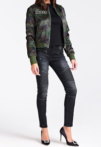Camo bomberjack met geribde manchetten en kraag, gecombineerd met strakke zwarte jeans met geribde details en zwarte stilettohakken met versiering.