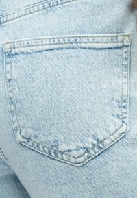 Svetlo modre jeans hlače iz denima z zadnjim žepom, ki ima trikotno polkrožno zaklopko, šivane robove in bledo teksturo z nežnimi gubami.