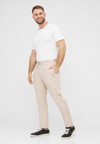Beige Jogginghose mit Taschen, schmaler Schnitt und dezentem Logo-Detail, kombiniert mit einem schlichten weißen T-Shirt und schwarzen Sneakers.