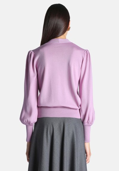 Femme aux longs cheveux foncés portant un pull violet clair à manches bouffantes et une jupe grise, vue de dos.