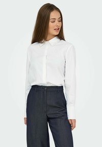 Chemise blanche à boutons avec des accents en perles sur le col, associée à un pantalon gris foncé ajusté, en coupe classique et en tissu texturé.