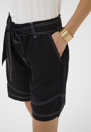 Persoon die een zwarte korte broek met witte stiksels en een gestrikte riem draagt, hand in de zak, en een brede gouden manchetarmband om de pols.