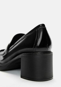 Schwarze Lackleder-Loafer mit einem klobigen Blockabsatz und sichtbaren Nähten an der Ferse vor einem weißen Hintergrund.