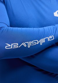 Άτομο που φορά μπλε πουκάμισο με άσπρο στυλιζαρισμένο κείμενο τυπωμένο στον πήχη, με σταυρωμένα τα χέρια.