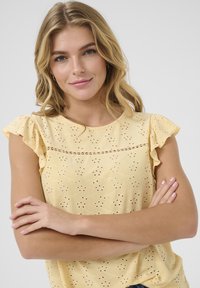 Blouse jaune à manches courtes volantes, ornée de broderie ajourée et d'une couture décorative horizontale. Tissu doux et léger.