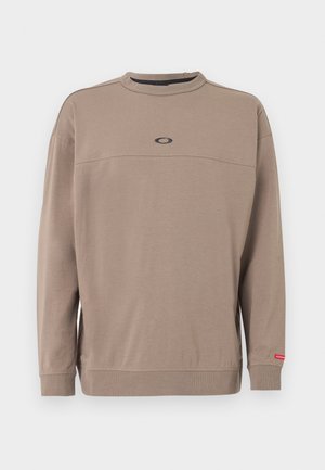 Langermet taupe-genser med rund hals, med en liten svart oval logo på brystet og en rød etikett på venstre mansjett.