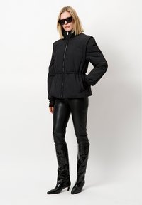 Zwarte pufferjack met een hoge kraag, elastische taille en ritssluiting; gecombineerd met aansluitende zwarte leren broeken en glanzende knielange laarzen.