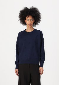 Mørkeblå strikkegenser med strukturmønster, rund hals, senkede skuldre og ribbestrikkede ermer og fald. Glatt overflate, avslappet passform.