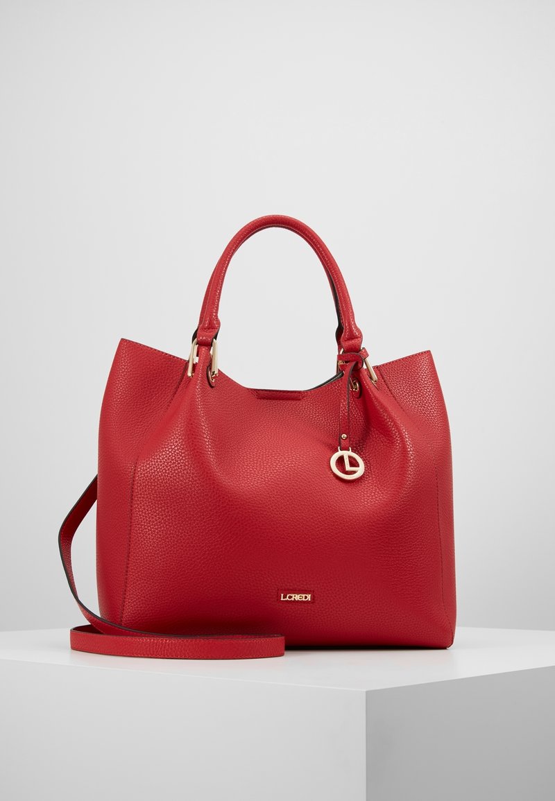 Sac à main en cuir texturé rouge avec deux poignées, une bandoulière amovible et un charm logo doré suspendu à une poignée.