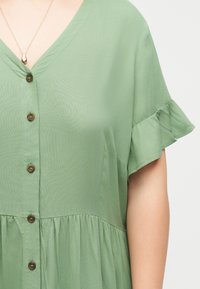 Γυναίκα με φόρεμα πράσινης φασκόμηλου με κουμπιά, κοντά βολάν μανίκια και κολιέ με δάκρυ κρεμαστό.