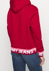 Sudadera con capucha roja y un ancho dobladillo rosa con la marca que dice "TOMMY JEANS". El material parece suave, presenta un corte holgado y un diseño en la parte trasera.