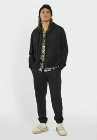 Casaco preto de botão sobre uma camisa floral verde, calças pretas e ténis de cor clara com solas texturizadas. O modelo tem cabelo longo.