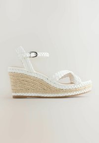 Sandale compensée en cuir blanc tressé avec sangle à la cheville et semelle plateforme en jute naturel tissé sur fond clair.