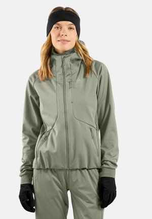 Frau trägt eine salbeigrüne Zip-Up-Jacke und passende Hose, schwarze Handschuhe und ein schwarzes Stirnband, steht mit Blick nach vorne.