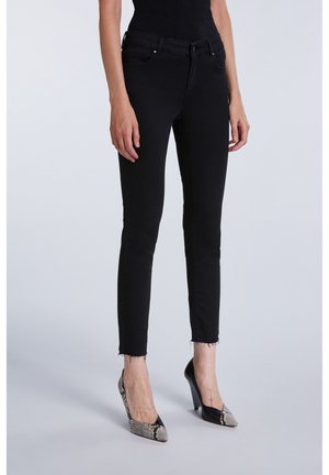 Vrouw die strakke zwarte spijkerbroeken draagt met onafgewerkte zomen en zwart-wit hoge hakken met slangenhuidpatroon, staand tegen een effen achtergrond.