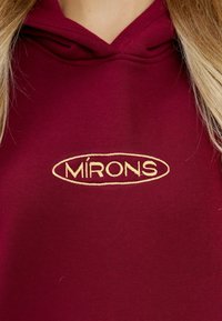 Felpa bordeaux con un tessuto testurizzato, caratterizzata da un ampio logo ricamato "MÍRONS" in oro, posizionato al centro della zona del petto.