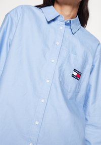 Light blue, long-sleeve button-up shirt made of cotton, featuring a front pocket with a multi-colored "Tommy Jeans" logo.