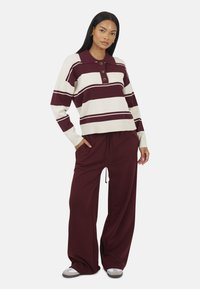 Langsleeve gestreepte trui in bordeauxrood en crème, met een kraag, drie knopen en een ontspannen pasvorm. Gecombineerd met bijpassende bordeauxrode broek.