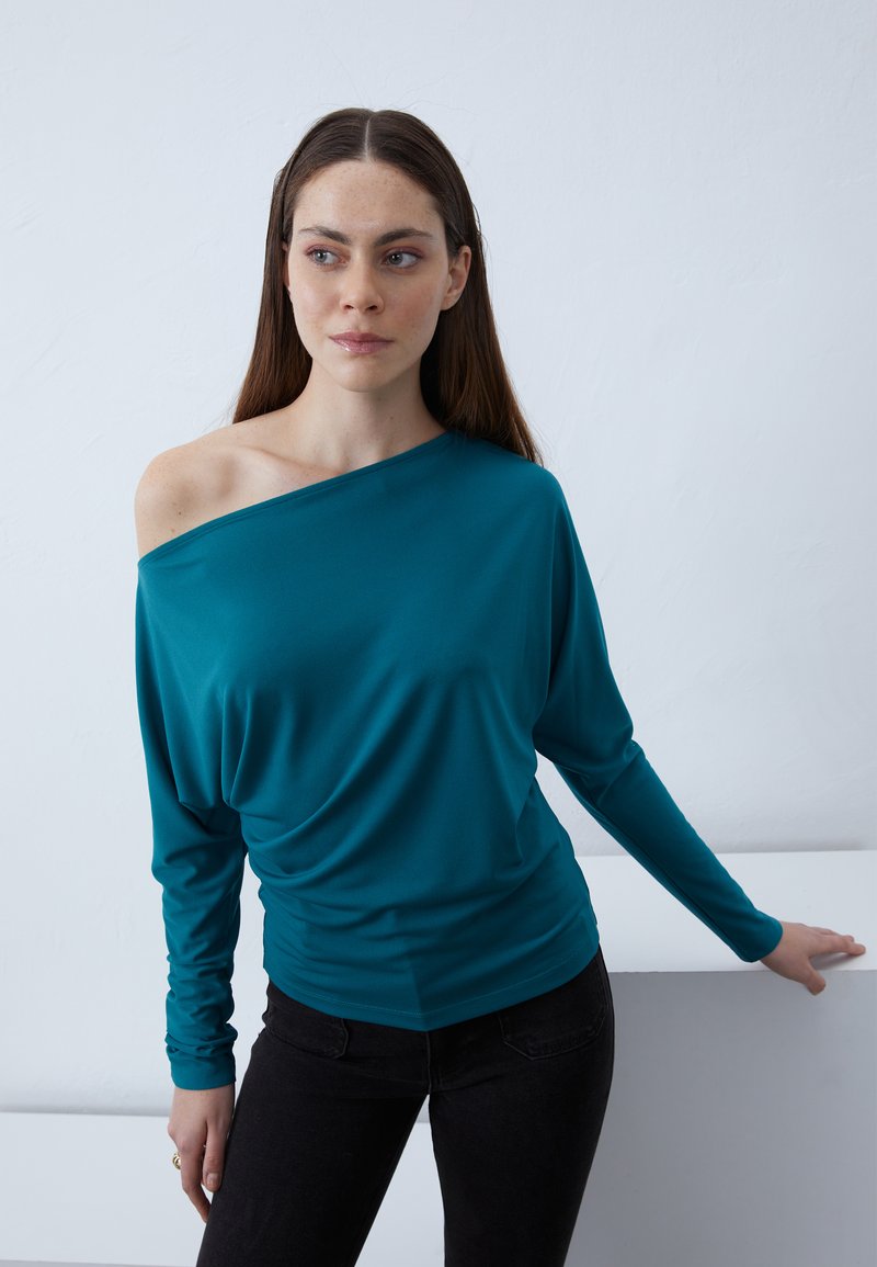 Zilganas off-shoulder tops ar garām piedurknēm, izgatavots no mīksta auduma; iekļauj drapētu dizainu un pieguļošu siluetu. Izskatu papildina melnas bikses.