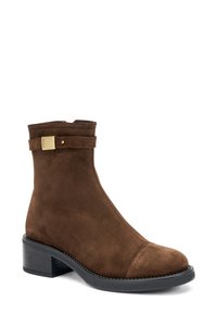 Stivale alla caviglia in suede marrone con tacco a blocco, presenta una cintura decorativa e hardware in tonalità oro. Tessuto liscio e design della punta rotonda.