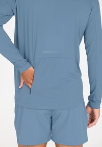 Långärmad, ljusblå träningsskjorta med en ficka med dragkedja och texturerat tyg, tillsammans med matchande shorts, sett från baksidan.