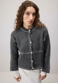 Mlada ženska s kodrasto rjavo lasi nosi temno sivo volneno kardigan s gumbi in belimi šivi čez belo krilo.