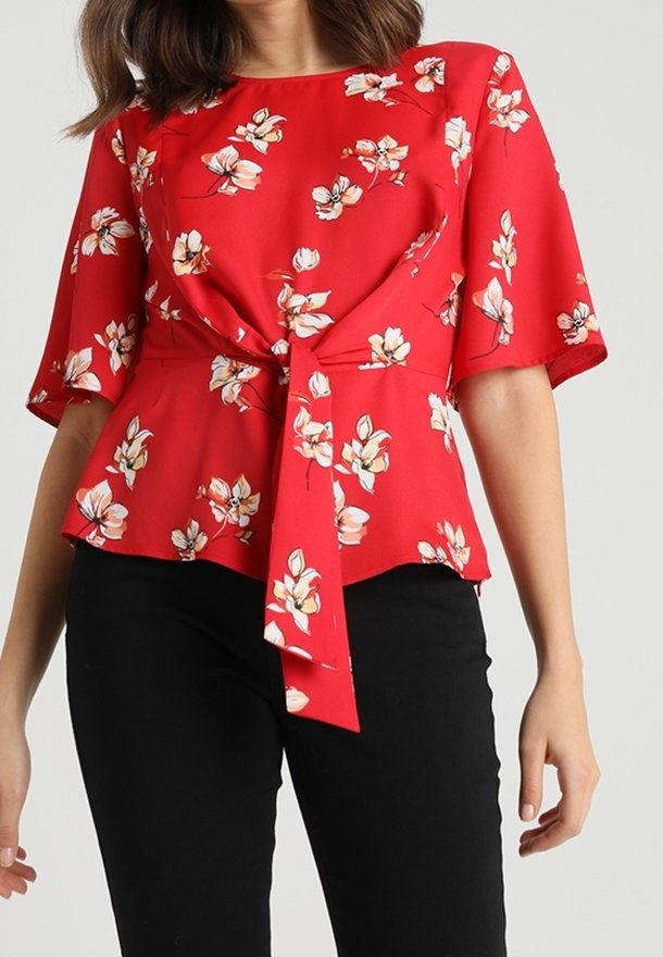 Femme portant un chemisier rouge à fleurs avec des manches aux coudes, un nœud à l'avant et un pantalon noir sur un fond clair uni.