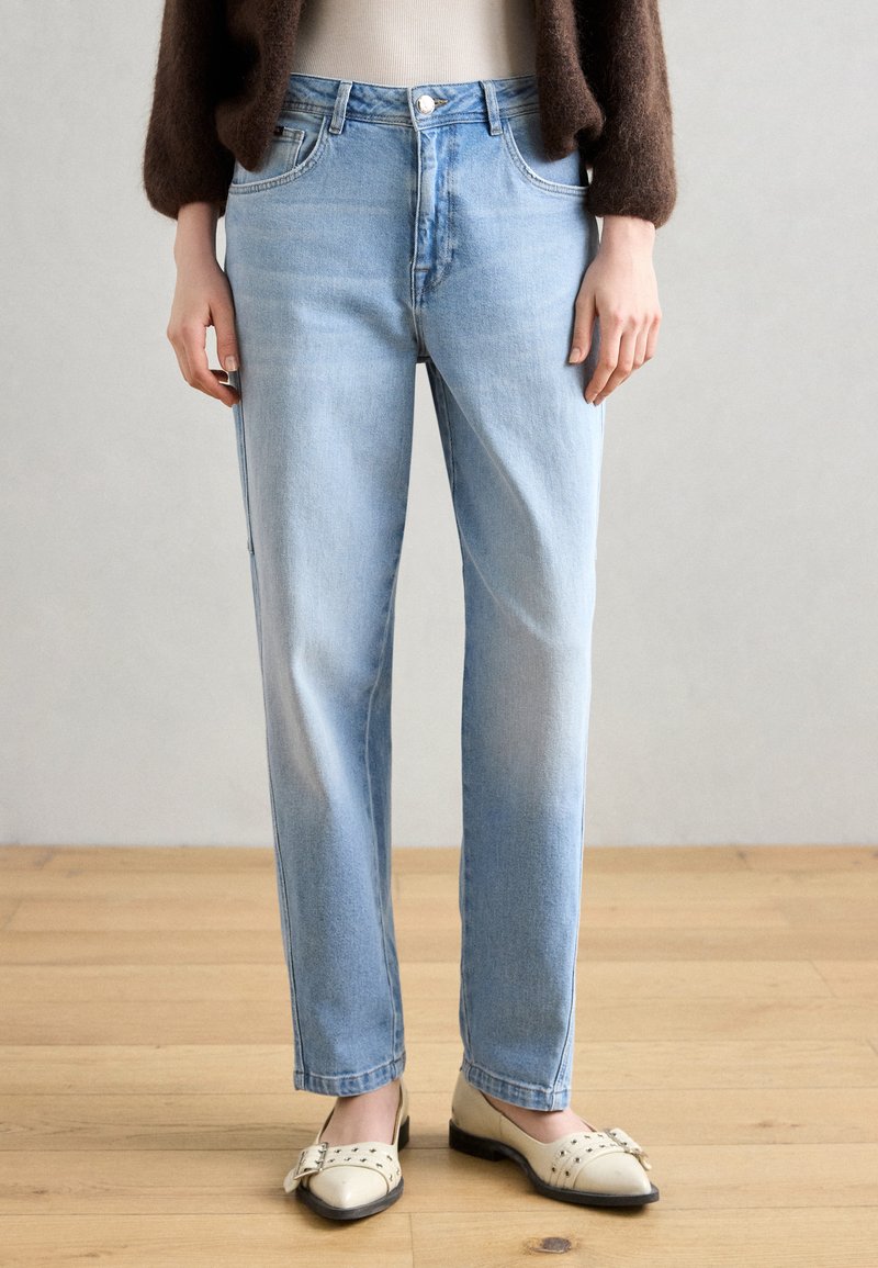Helle blaue, gerade geschnittene Jeans aus Denim mit verblassten Akzenten, fünf Taschen und einem einzelnen Knopfverschluss, kombiniert mit cremefarbenen, flachen Schuhen.