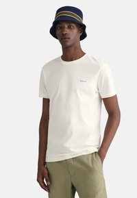 Hvid T-shirt med rund hals og korte ærmer, prydet med et lille blåt "GANT" logo på brystet. Bæret med lysegrønne linnedshorts og en blå bøttehue.