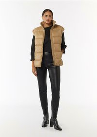 Beige Puffer-Weste mit hohem Kragen, ärmellosem Design, getragen über einem schwarzen Langarmoberteil, kombiniert mit schwarzen Lederhosen und Stiefeln mit Absatz.