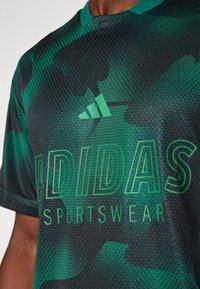 Zeleno-černé sportovní tričko s krátkým rukávem a texturovanou tkaninou, s výrazným logem Adidas a nápisem "SPORTWEAR" tučně.