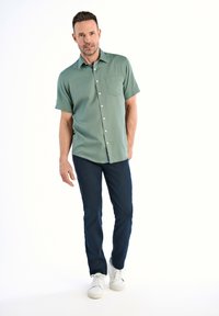 Camicia verde a maniche corte con bottoni e taschino, abbinata a jeans blu scuro e sneakers bianche. Sfondo neutro.