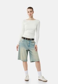 Hvid langærmet top, lyseblå denimbermuda-shorts med vaskede detaljer, sort bælte, hvide sokker og beige sneakers. Enkle runde øreringe.