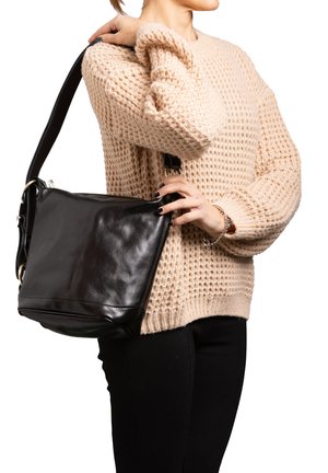 Frau in beige gestricktem Pullover hält schwarze Leder-Schultertasche mit verstellbarem Riemen vor weißem Hintergrund.