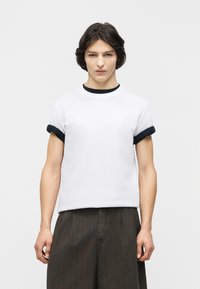 Νεαρό άτομο με σκούρα μαλλιά που φοράει λευκό μπλουζάκι με μαύρη λεπτομέρεια και φαρδιά σκοτεινά παντελόνια με ρίγες, στέκεται μπροστά σε απλό φόντο.